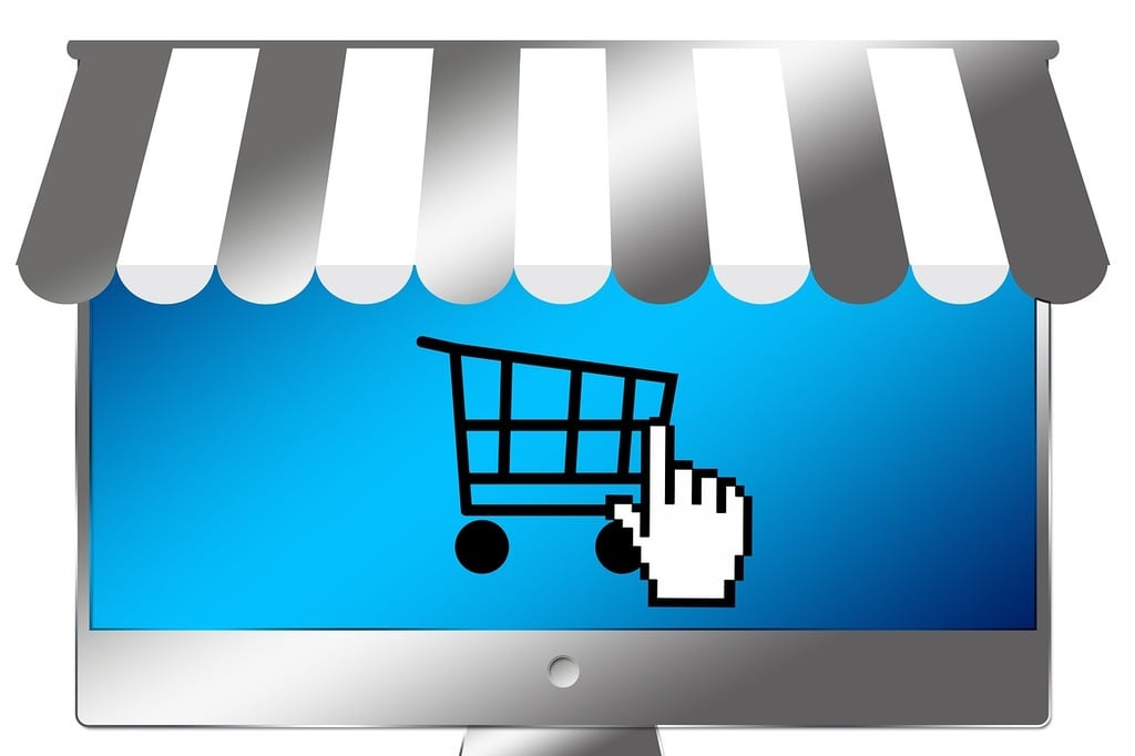 A stylized e-commerce storefront showing a computer screen with a shop awning. A pointer icon hovers over a shopping cart on the screen.