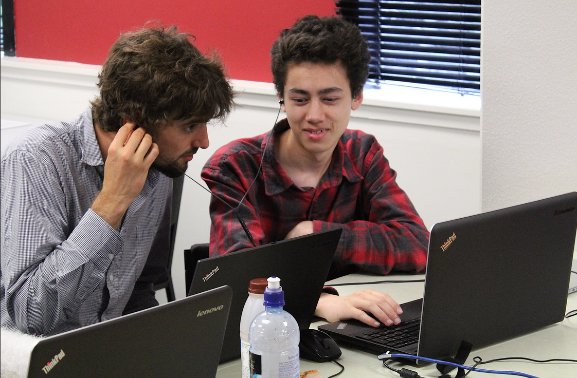 Two men sharing one set of earbuds while working on a laptop.